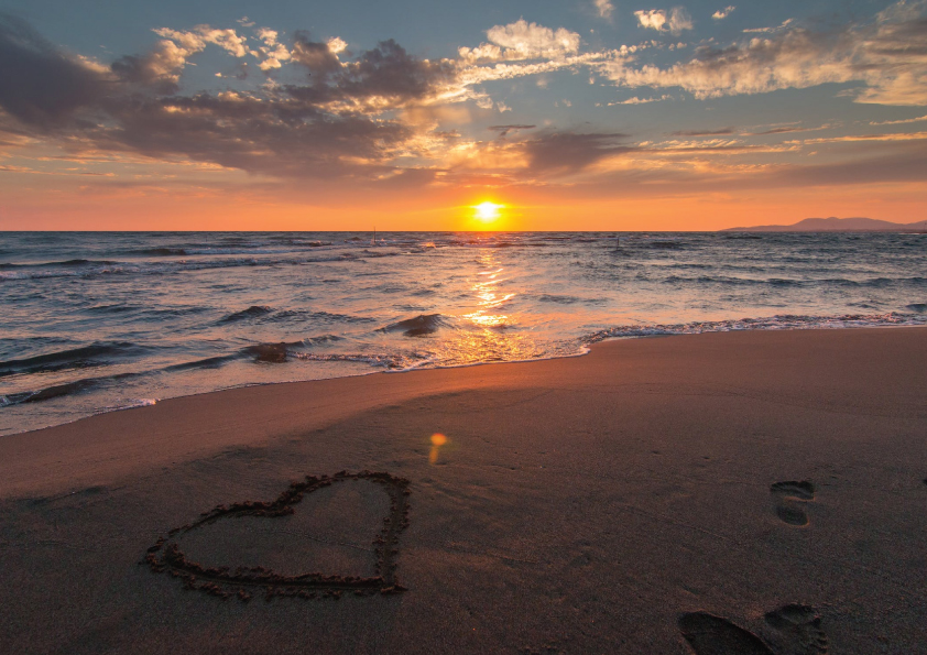 Widok plaży z sercem narysowanym na piasku, w tle zachodzące słońce.