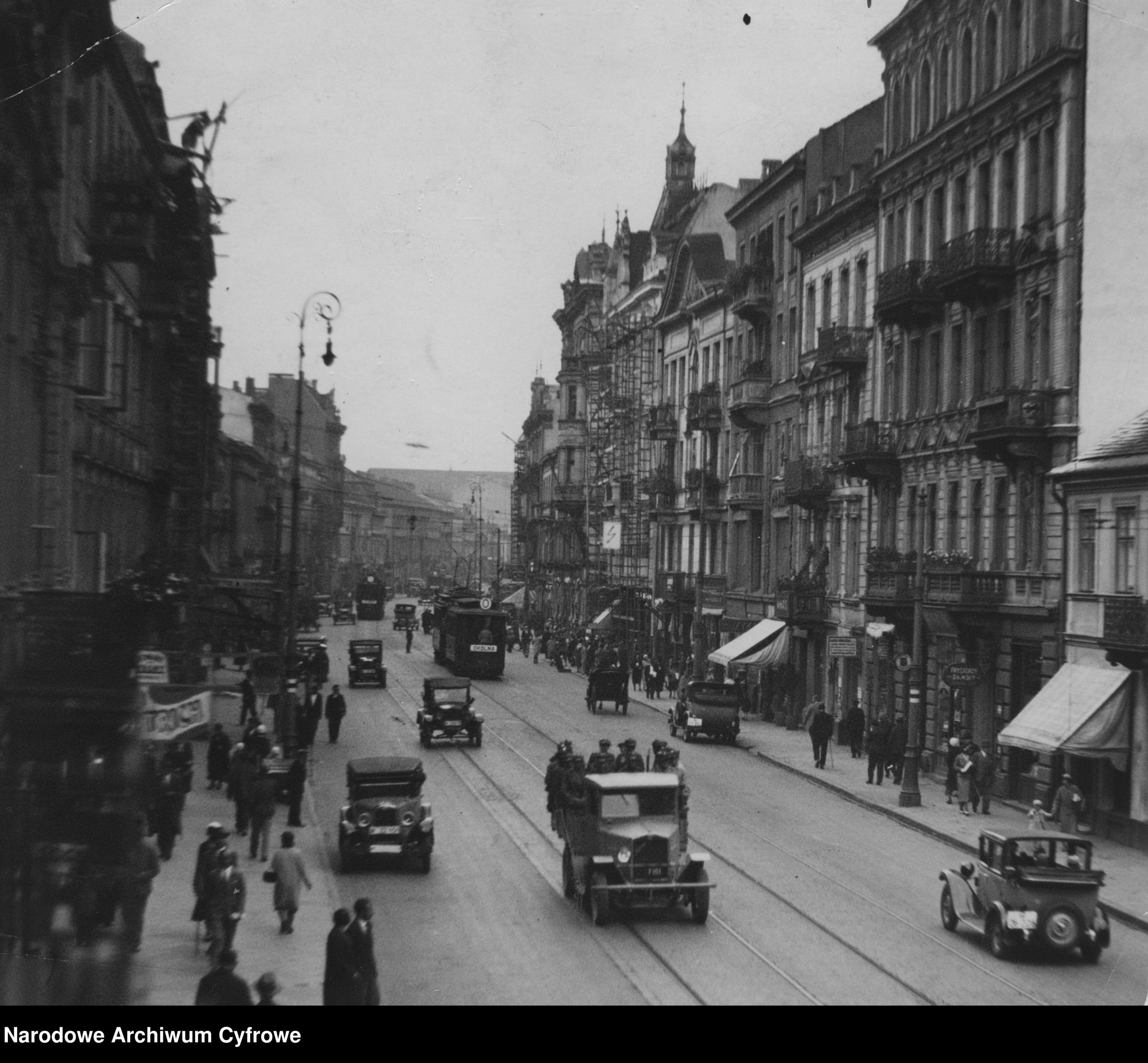 ulica z lat 30. widać samochody jadące po torowisku, wzdłuż kamienic idą ludzie.